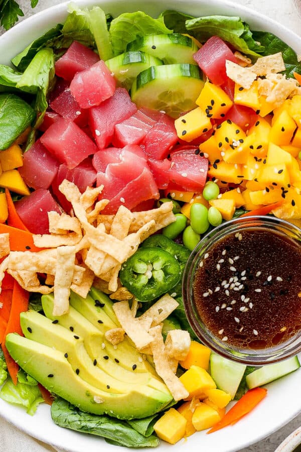 Top down shot of a poke salad in a shallow bowl with tuna, avocado, mango, carrot and a small bowl of sesame ginger dressing.