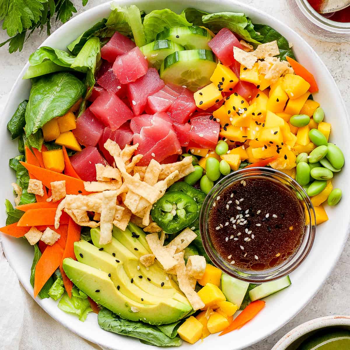Top down shot of a poke salad in a shallow bowl with tuna, avocado, mango, carrot and a small bowl of sesame ginger dressing.