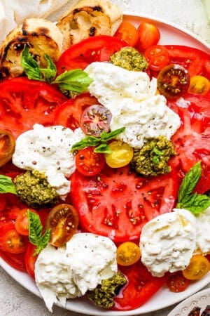 A large plate filled with sliced tomatoes, basil, pesto, burrata cheese and some grilled bread slices on the side.