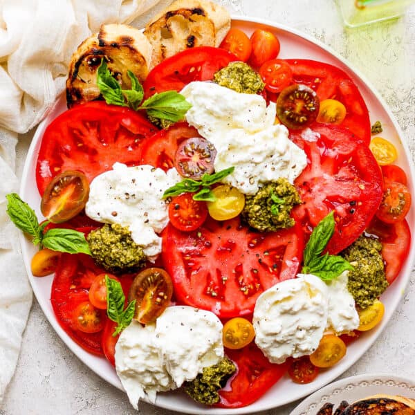 A large plate filled with sliced tomatoes, basil, pesto, burrata cheese and some grilled bread slices on the side.