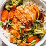 Top down shot of a bowl of rice with baked teriyaki chicken and veggies on top with sesame seed garnish.