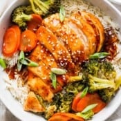 Top down shot of a bowl of rice with baked teriyaki chicken and veggies on top with sesame seed garnish.