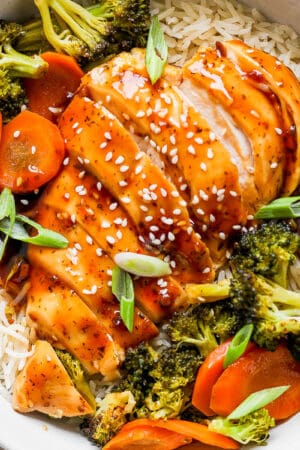 Top down shot of a bowl of rice with baked teriyaki chicken and veggies on top with sesame seed garnish.