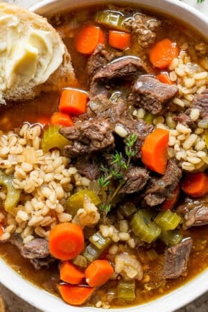 Top down shot of a bowl of beef and barley soup with a piece of bread sticking out of it.