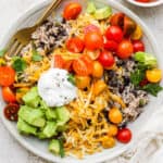 Top down shot of a plate filled with black beans and rice with shredded cheese, tomato, avocado and sour cream on top.