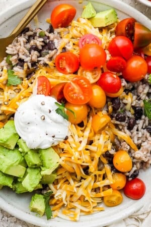 Top down shot of a plate filled with black beans and rice with shredded cheese, tomato, avocado and sour cream on top.