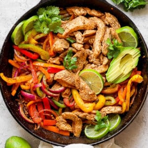 Top down shot of a cast iron skillet filled with chicken fajitas with lime slices, avocado and cilantro.