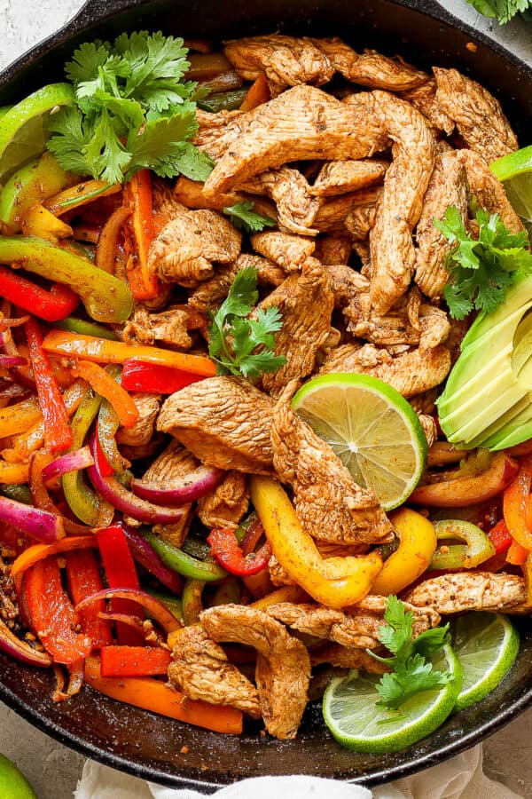 Top down shot of a cast iron skillet filled with chicken fajitas with lime slices, avocado and cilantro.