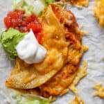 A crispy chicken taco on a piece of parchment paper with melted cheese, salsa, guacamole and sour cream on top.