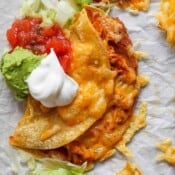 A crispy chicken taco on a piece of parchment paper with melted cheese, salsa, guacamole and sour cream on top.