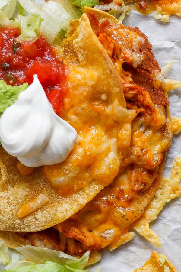 A crispy chicken taco on a piece of parchment paper with melted cheese, salsa, guacamole and sour cream on top.