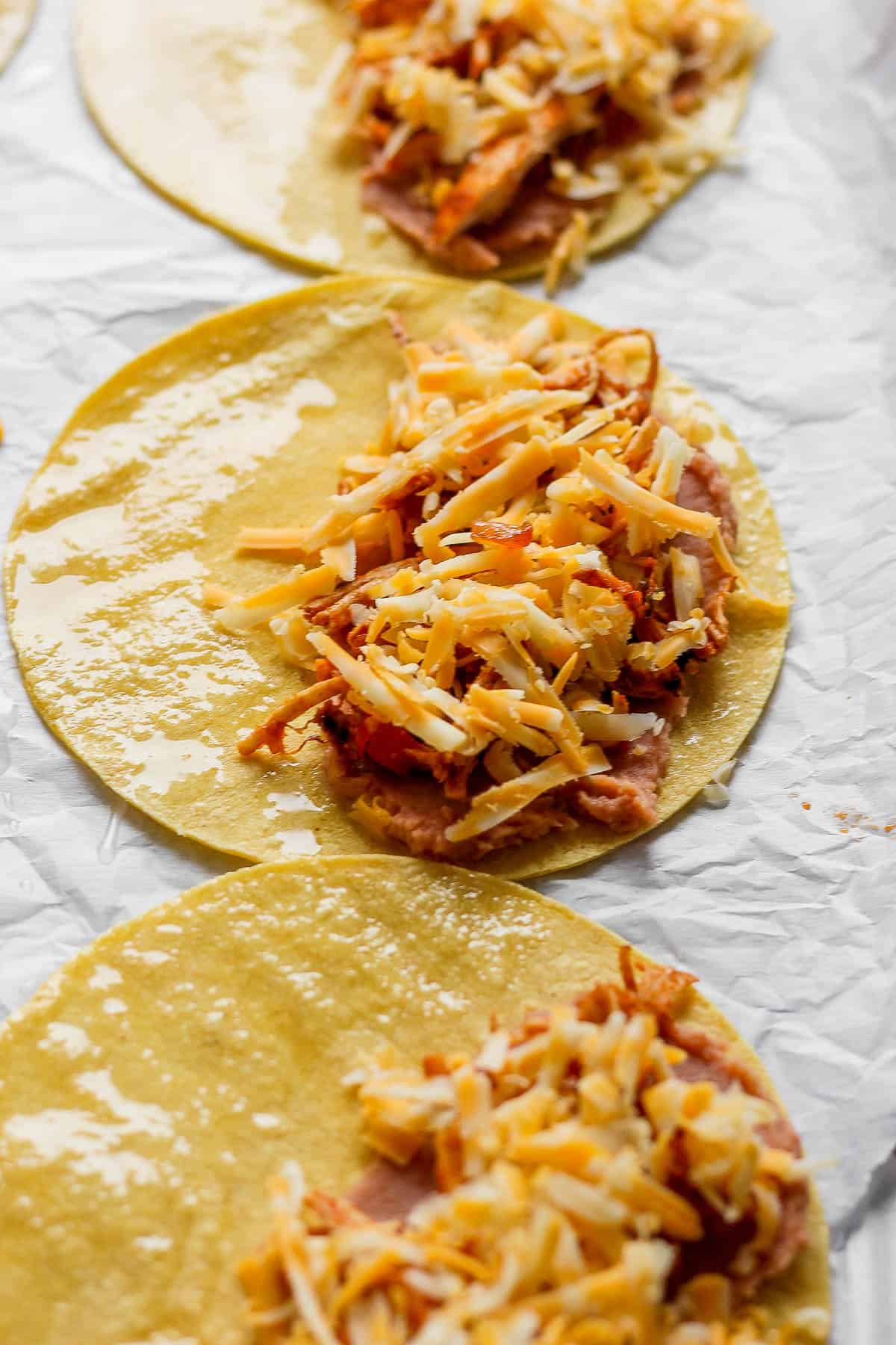 Tortillas on a parchment-lined baking sheet with refried beans, chicken, and cheese on one side of them.