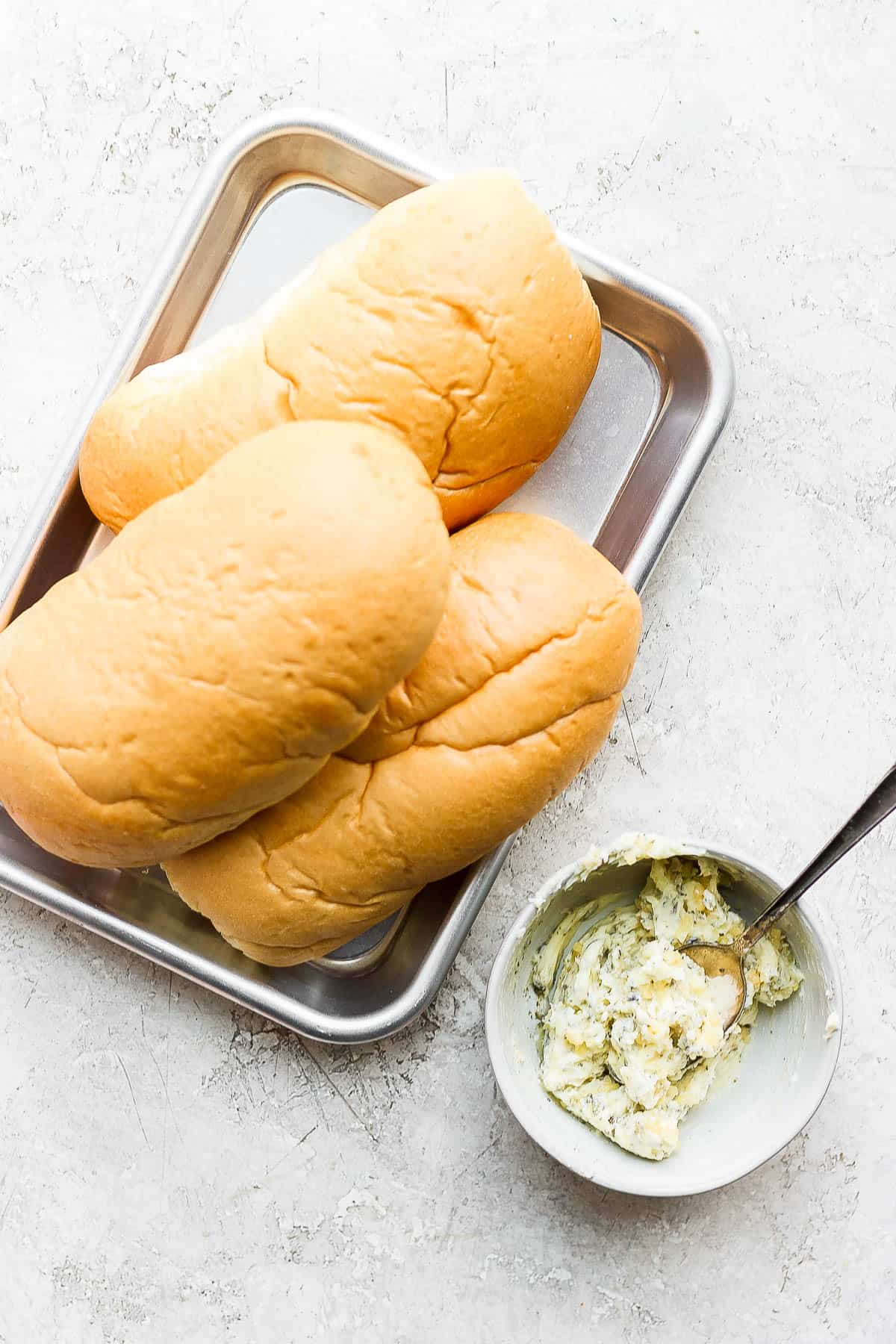 Three hoagie buns on a counter with a bowl of herbed butter on the side.