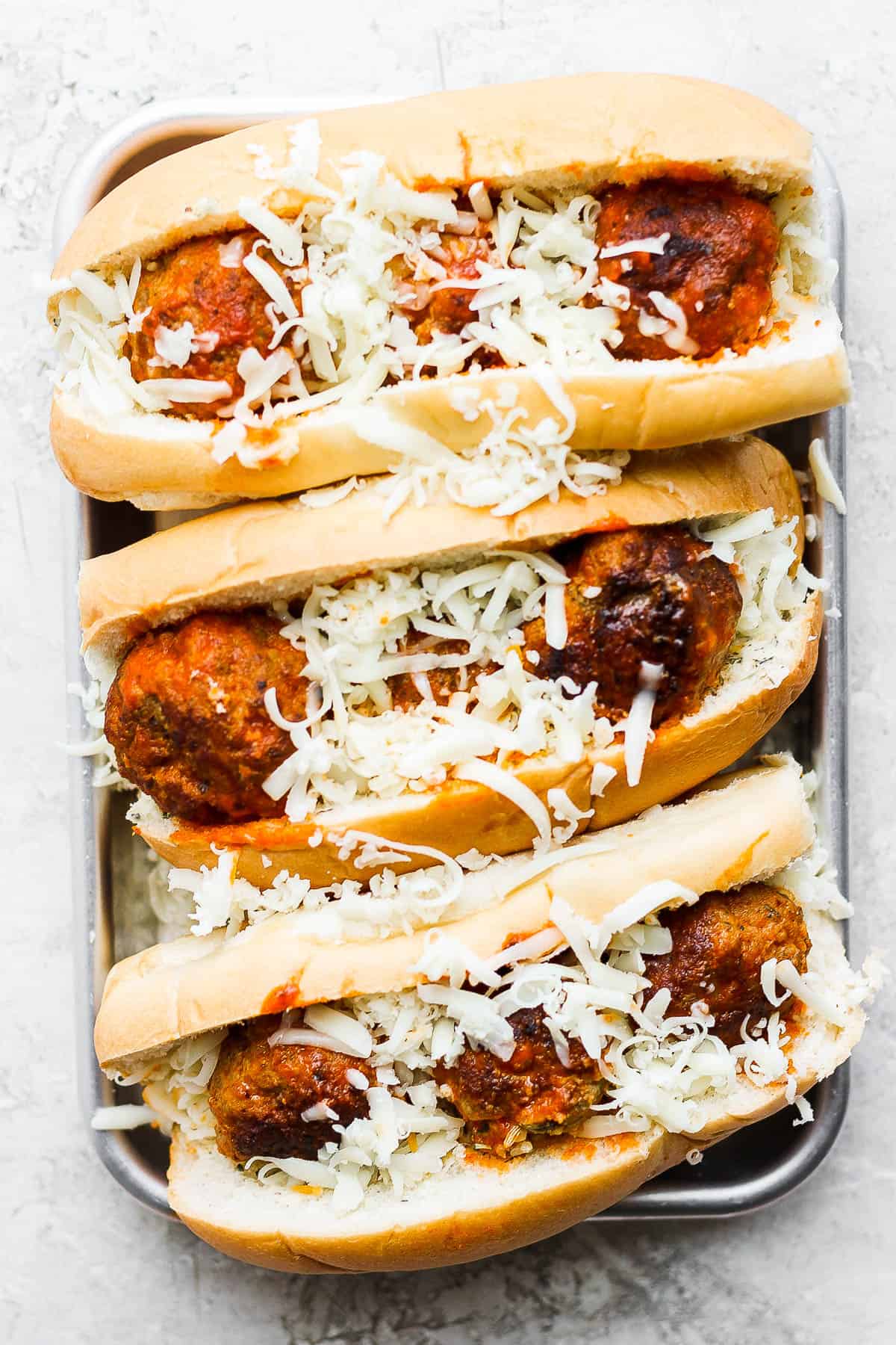 Three assembled meatball subs on a baking sheet sprinkled with mozzarella cheese.