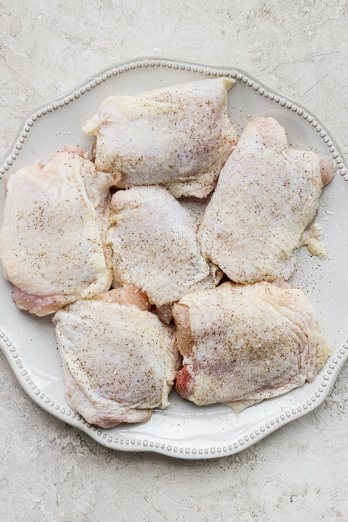 Raw chicken thighs seasoned with salt and pepper on a large plate.