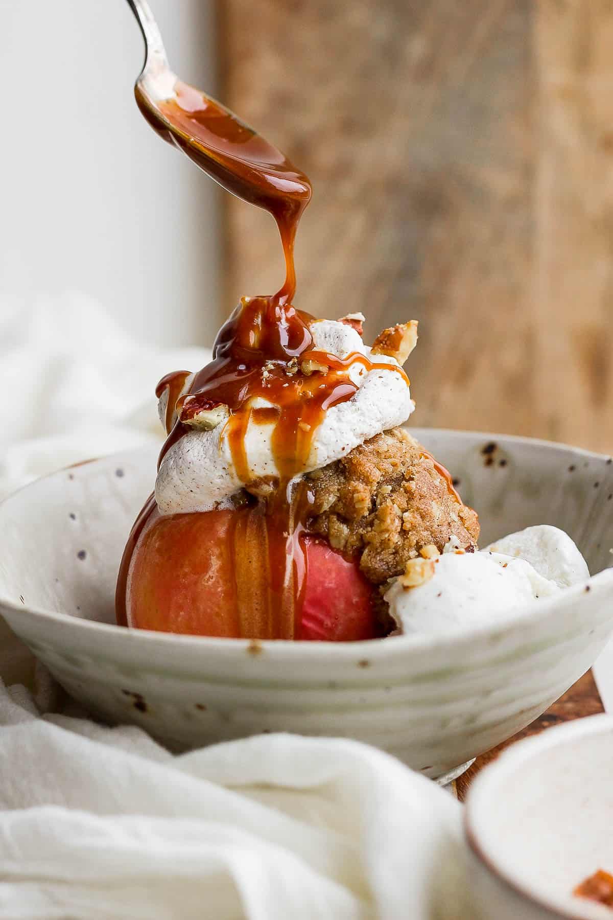 A baked apple in a bowl with ice cream, whipped cream, walnuts, and caramel being drizzled on top.