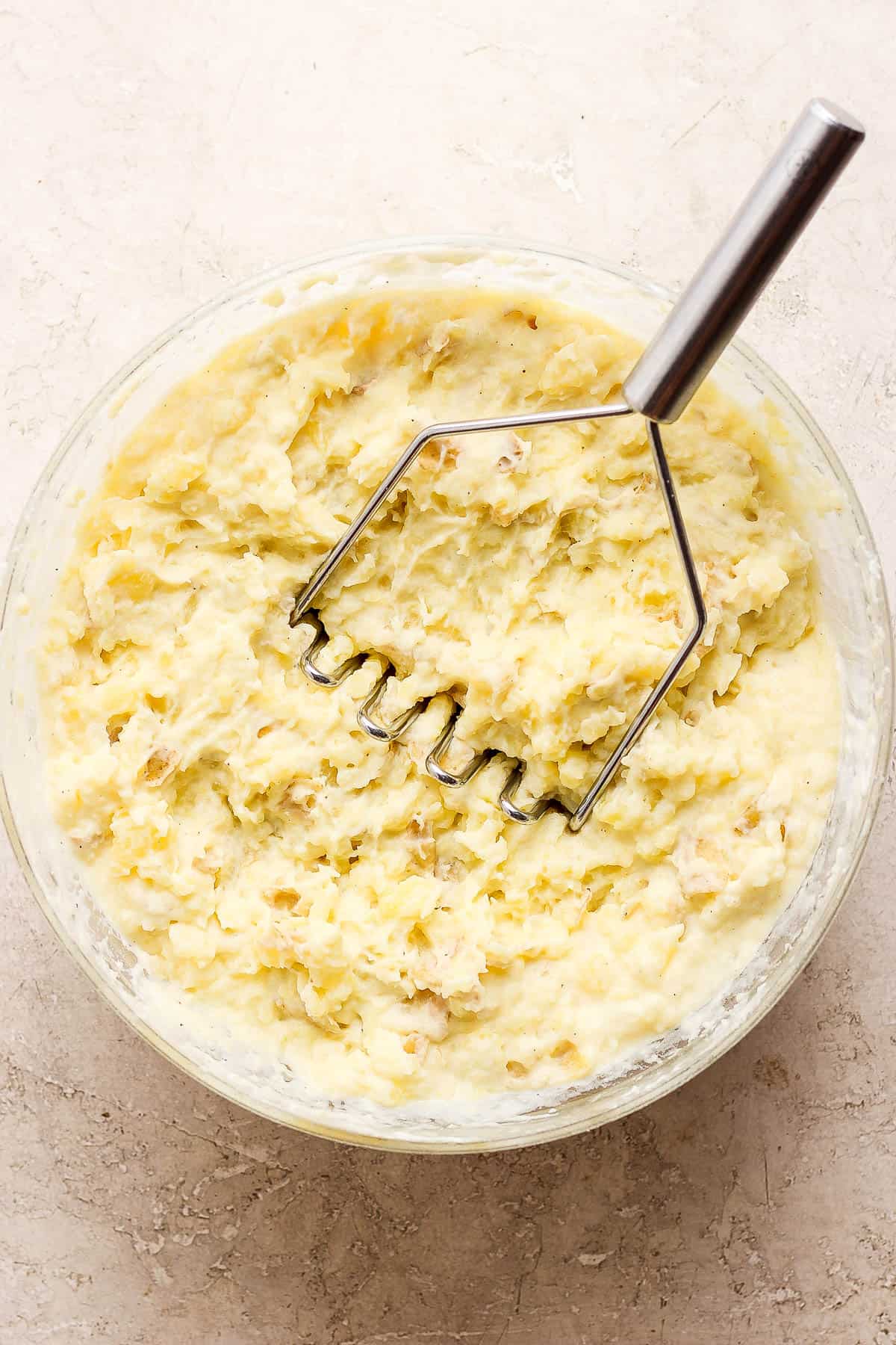 A potato masher in the bowl with the potatoes fully mashed.
