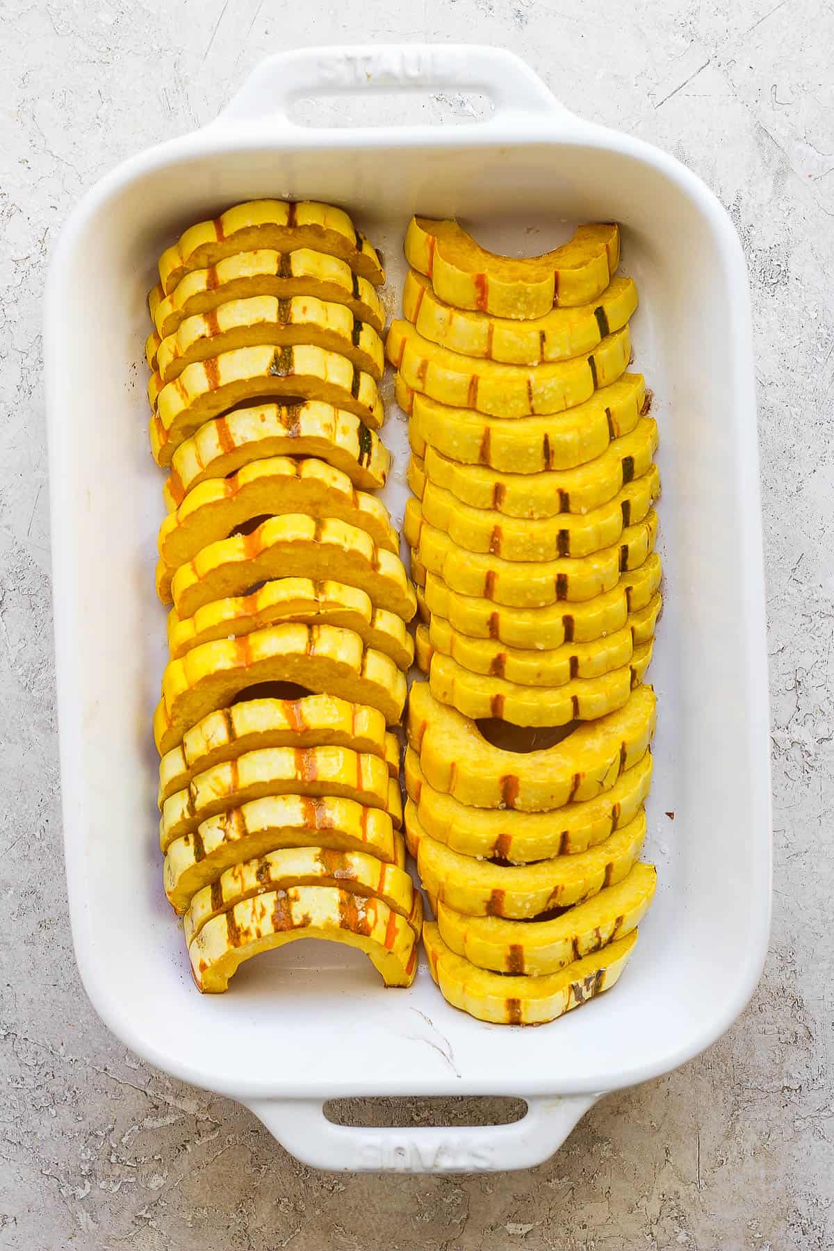 Sliced delicata squash in a baking dish after being tossed in oil and seasoned with salt and pepper.