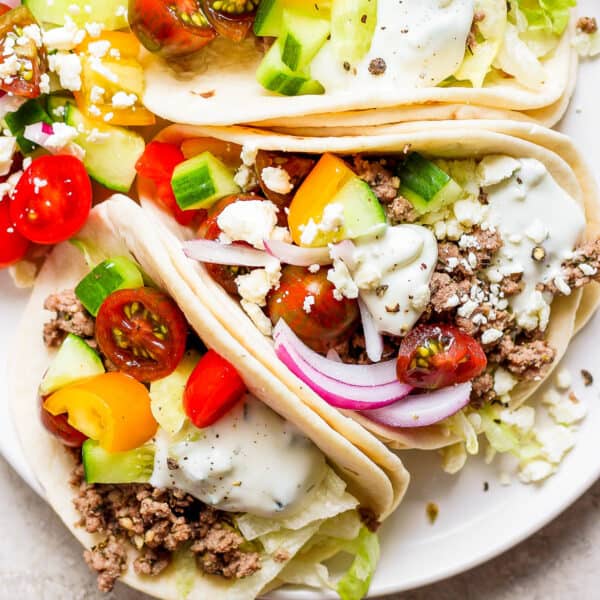 Top down shot of a plate of three lamb tacos with onion, cherry tomatoes, feta and tzatziki sauce.