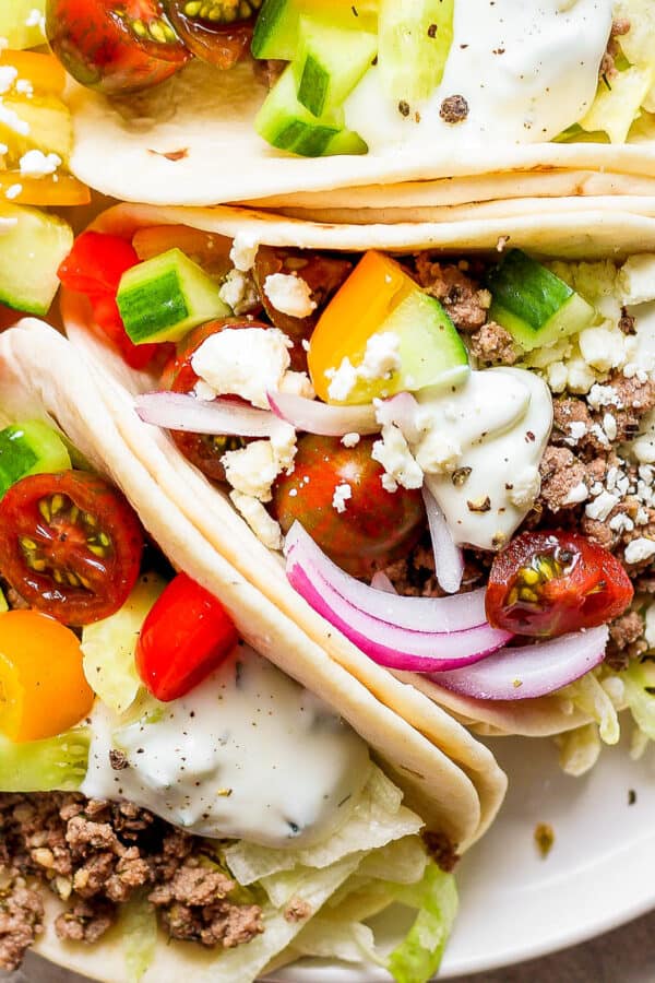 Top down shot of a plate of three lamb tacos with onion, cherry tomatoes, feta and tzatziki sauce.
