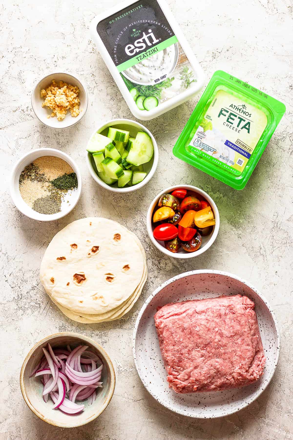 Individual bowls of cut cherry tomatoes, quartered cucumbers, sliced red onion, raw ground lamb, a stack of tortillas, a package of feta cheese, a bowl of seasonings, minced garlic, and tzatziki sauce.