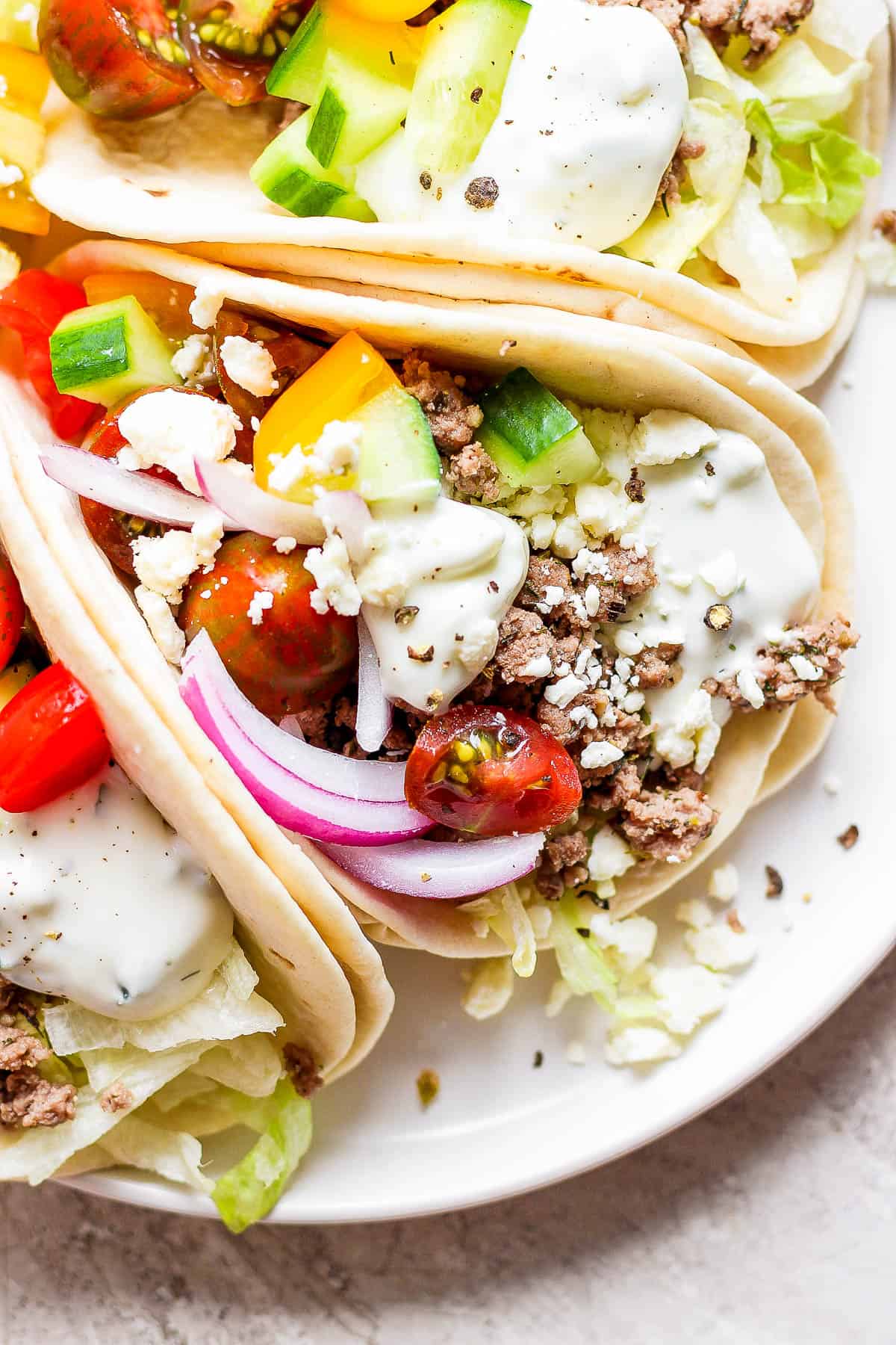Three lamb tacos on a plate topped with red onion, cucumber, cherry tomatoes, feta, and tzatziki sauce.