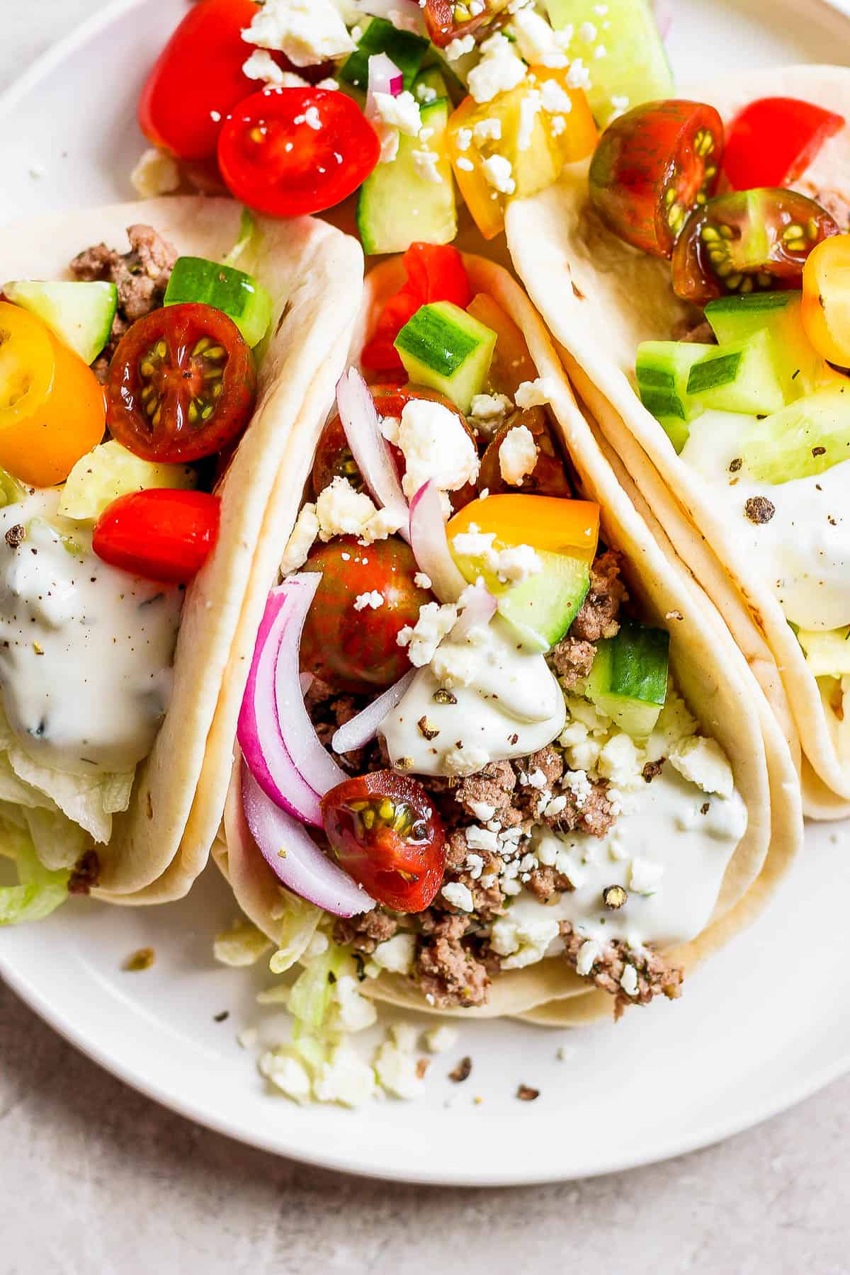 Three lamb tacos on a plate topped with red onion, cucumber, cherry tomatoes, feta, and tzatziki sauce.