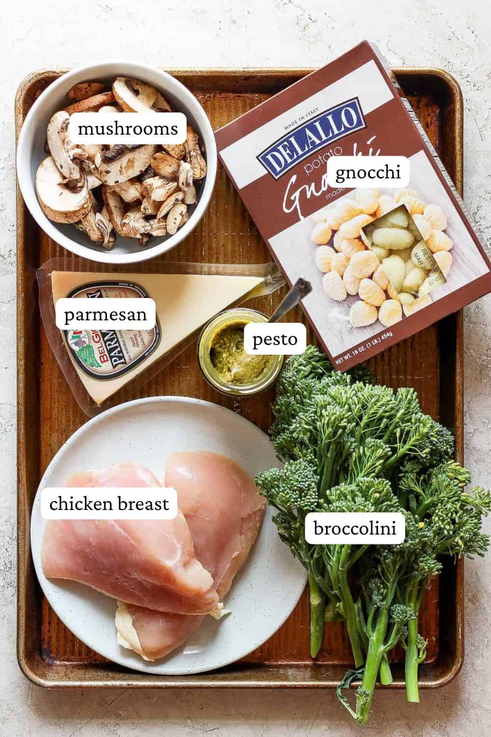 Sheet pan chicken and gnocchi ingredients on a sheet pan - sliced mushrooms, gnocchi, parmesan, pesto, chicken breasts, and broccolini.