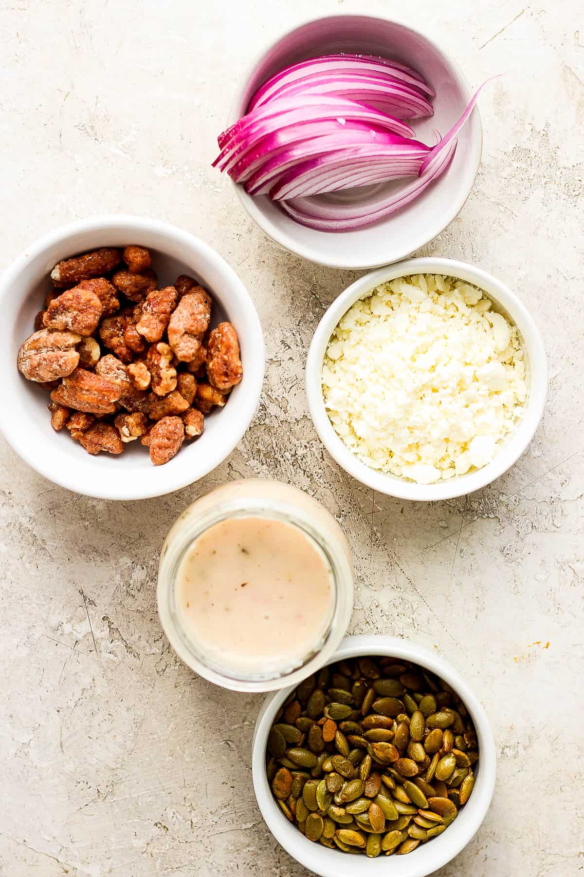Individual bowls of sliced onion, candied pecans, feta cheese, dressing, and pepitas.
