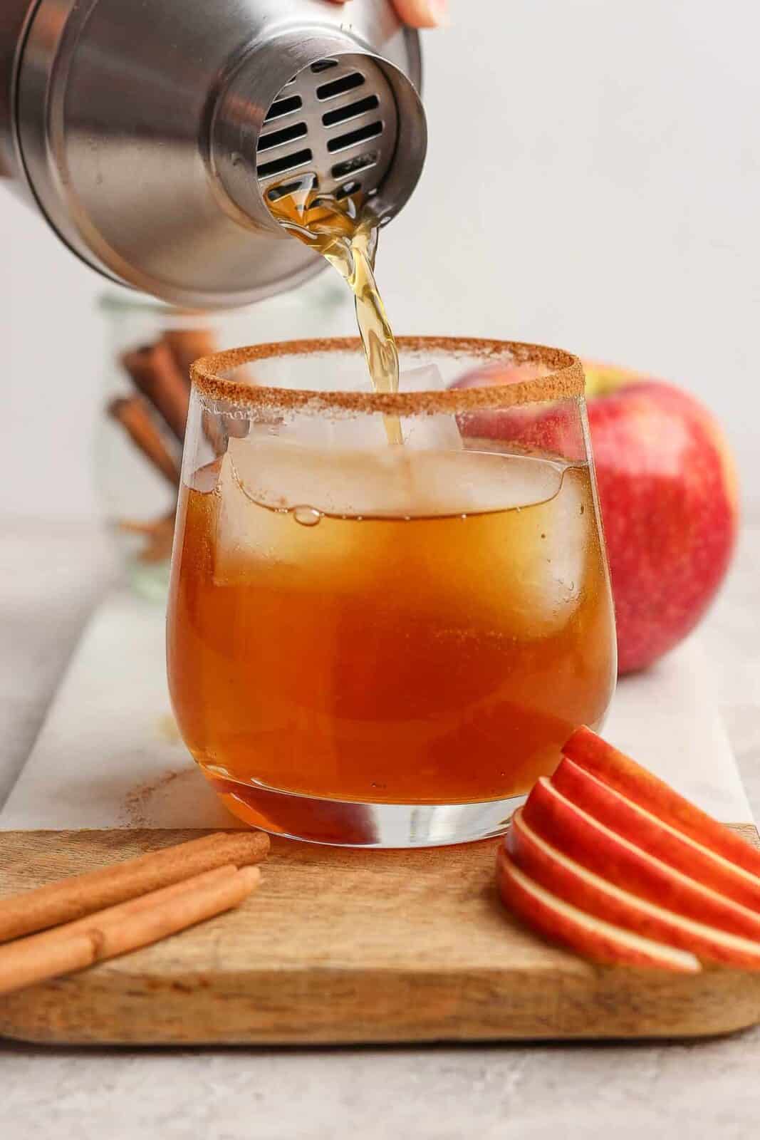 Someone pouring an apple cider margarita into a glass that has ice cubes in it and a cinnamon sugar rim with sliced apples and a cinnamon stick in front of it.