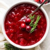 Top down shot of a bowl of classic homemade cranberry sauce with a spoon sticking out and three raw cranberries and two sprigs of rosemary for garnish.