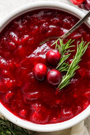 Top down shot of a bowl of classic homemade cranberry sauce with a spoon sticking out and three raw cranberries and two sprigs of rosemary for garnish.
