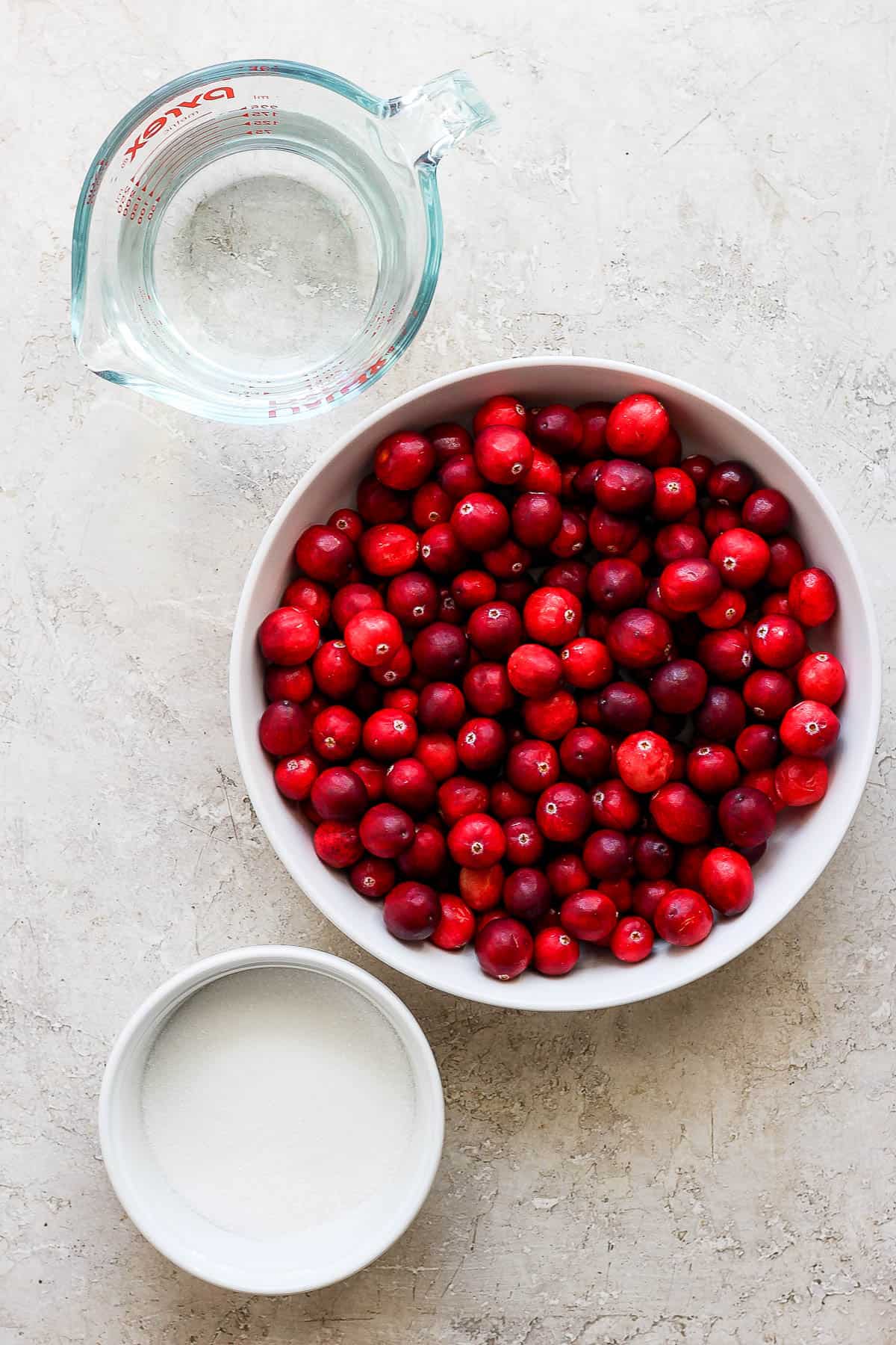 A bowl of fresh cranberries, a measuring cup with water, and a bowl of sugar.