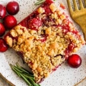 Top down shot of a cranberry bar on a plate with a fork, some raw cranberries and a few pieces of rosemary.