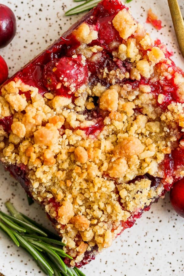 Top down shot of a cranberry bar on a plate with a fork, some raw cranberries and a few pieces of rosemary.