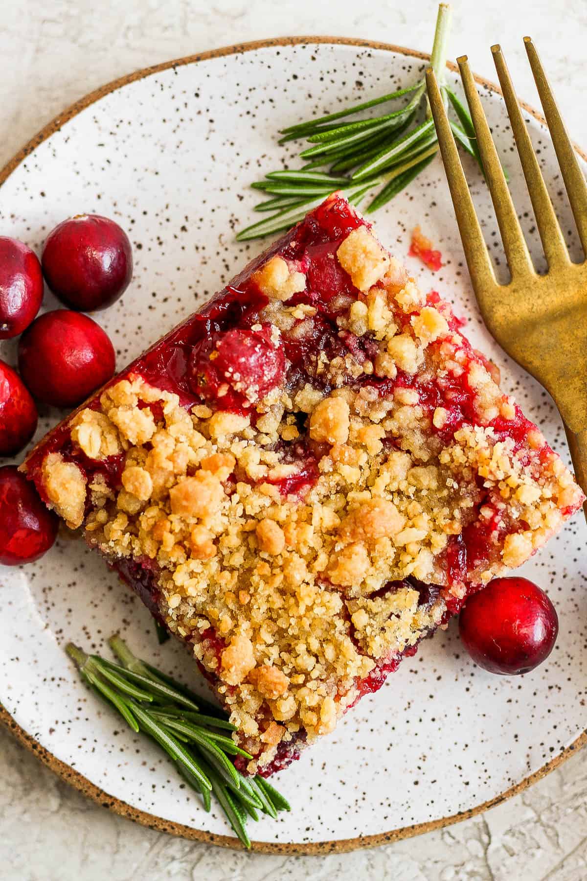 A cranberry bar on a plate alongside a fork, fresh rosemary, and fresh cranberries.