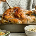 A dry brined turkey in a roasting pan with someone behind it brushing butter on it.
