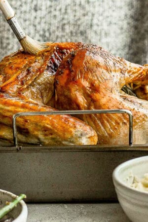 A dry brined turkey in a roasting pan with someone behind it brushing butter on it.