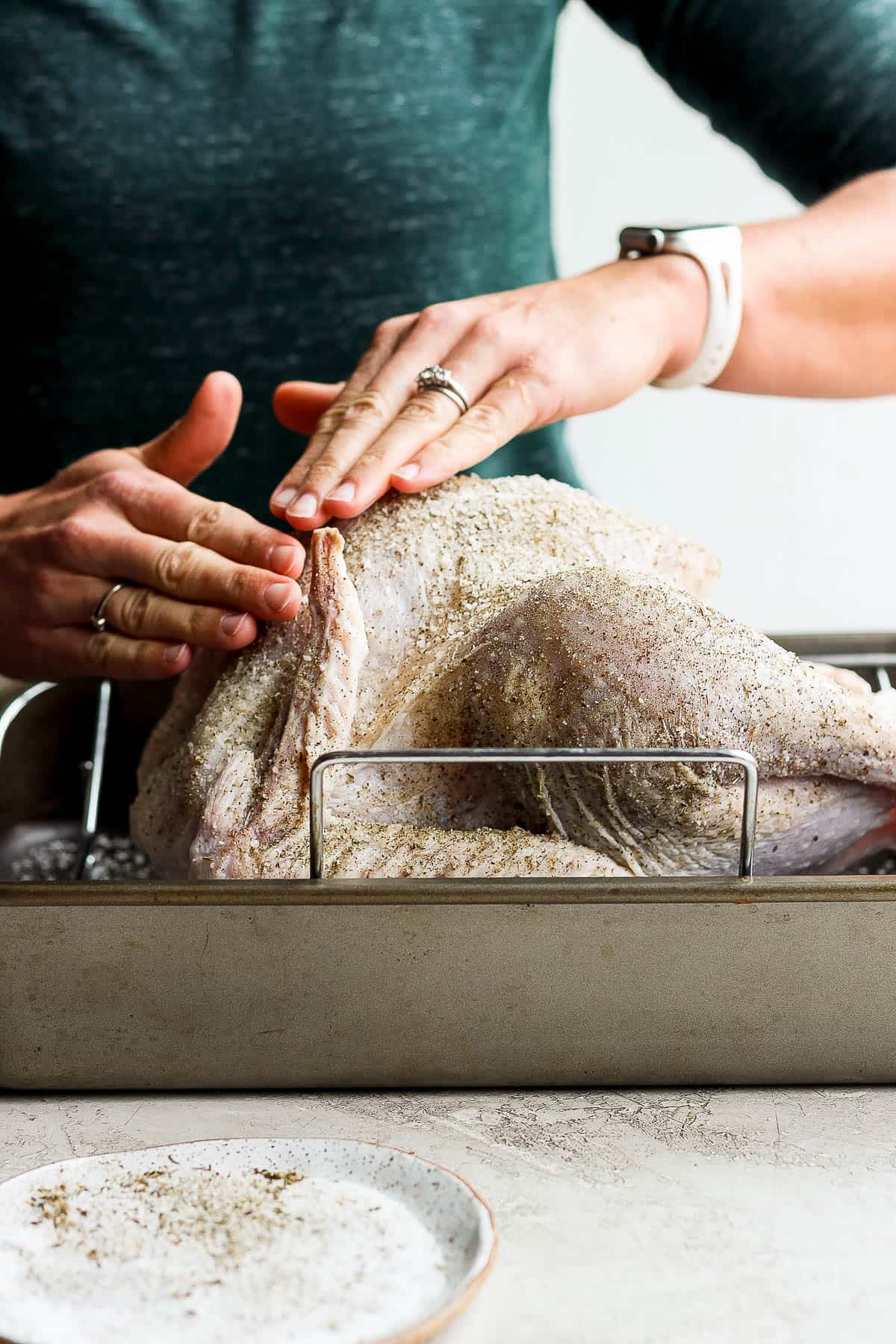 A whole turkey in a roasting pan being rubbed with a dry brine.