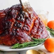 A grilled ham on a plate with fresh rosemary garnish and someone brushing on a ham glaze.