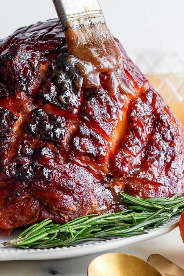 A grilled ham on a plate with fresh rosemary garnish and someone brushing on a ham glaze.