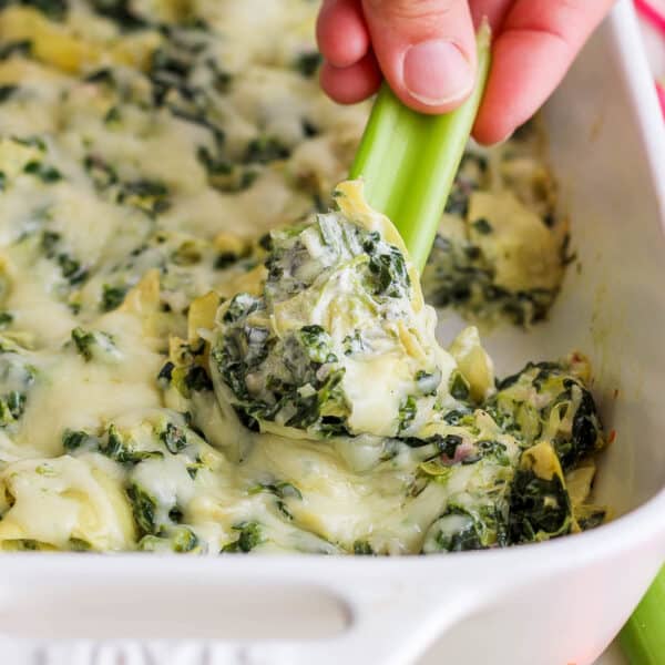 Someone using a piece of celery to scoop out some healthy spinach artichoke dip from a baking dish.