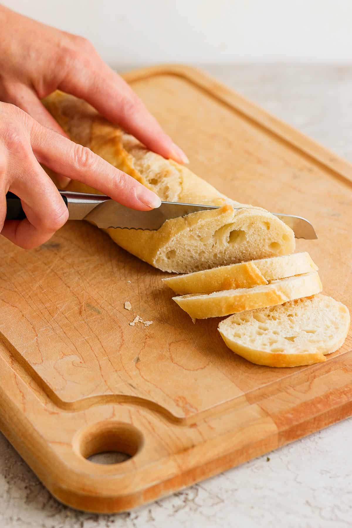 A french baguette being cut into slices on a cutting bread with a serrated knife.