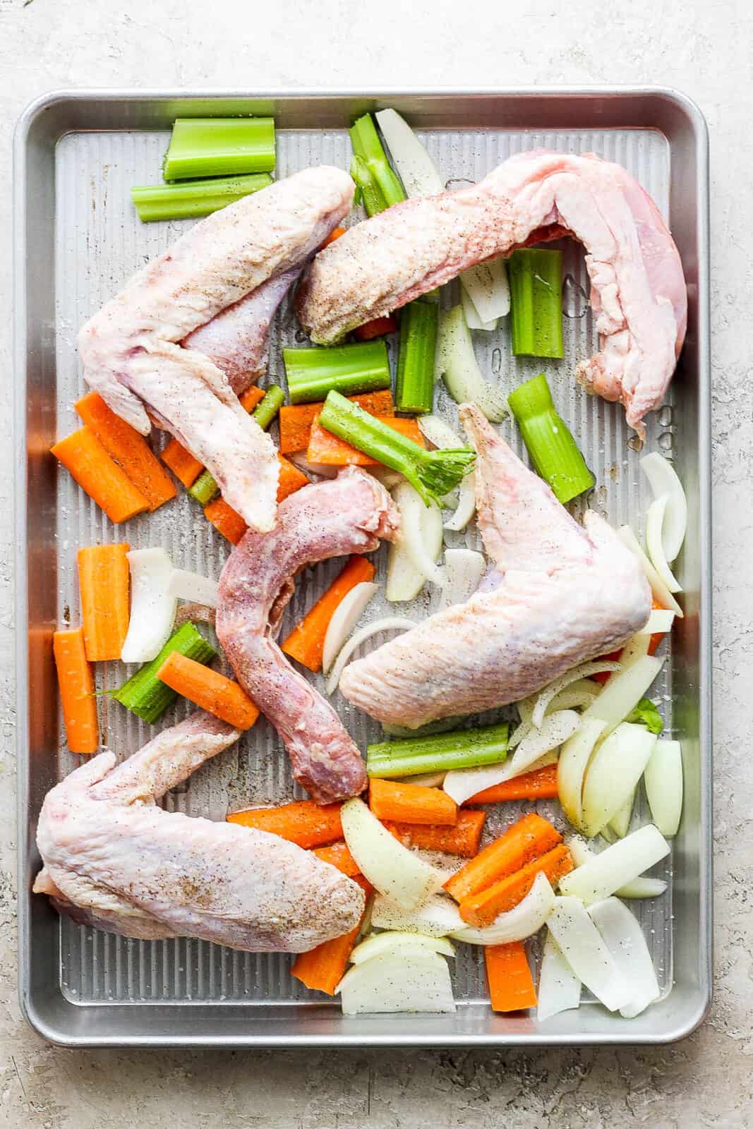 Raw turkey legs, carrots, celery, and onion on a baking sheet seasoned with olive oil and salt and pepper.