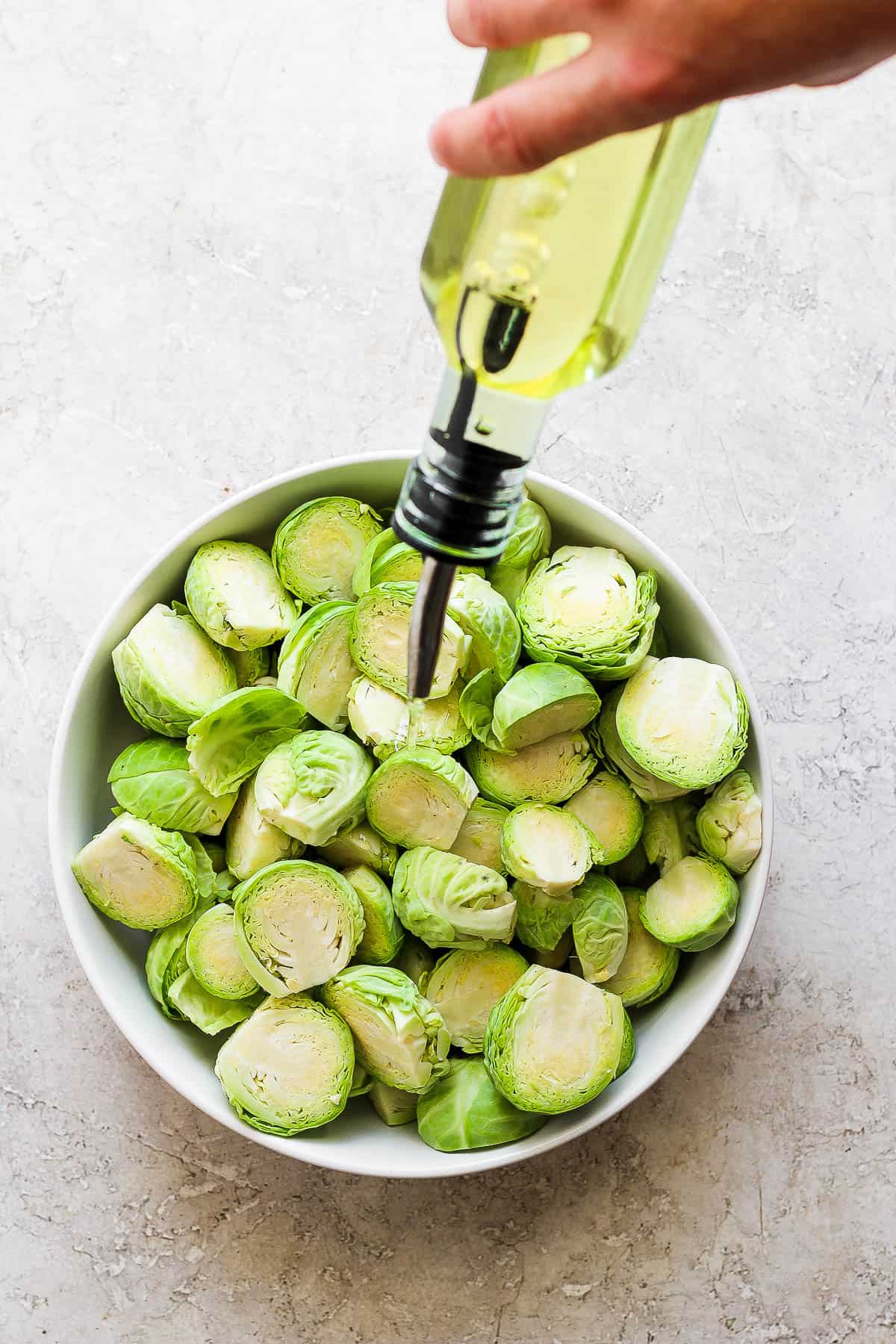 A bowl of trimmed and halved brusssel sprouts being drizzled with olive oil.