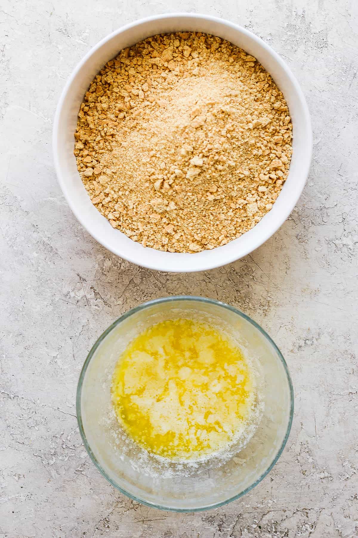 A white bowl with crushed graham crackers and a glass bowl with melted butter.