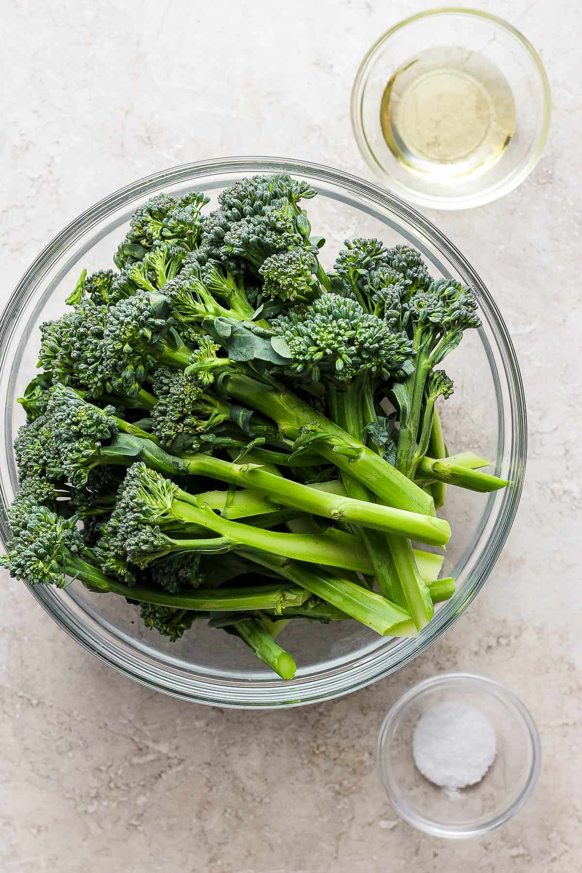 A large glass bowl of fresh broccolini and two smaller glass bowls of oil and salt.