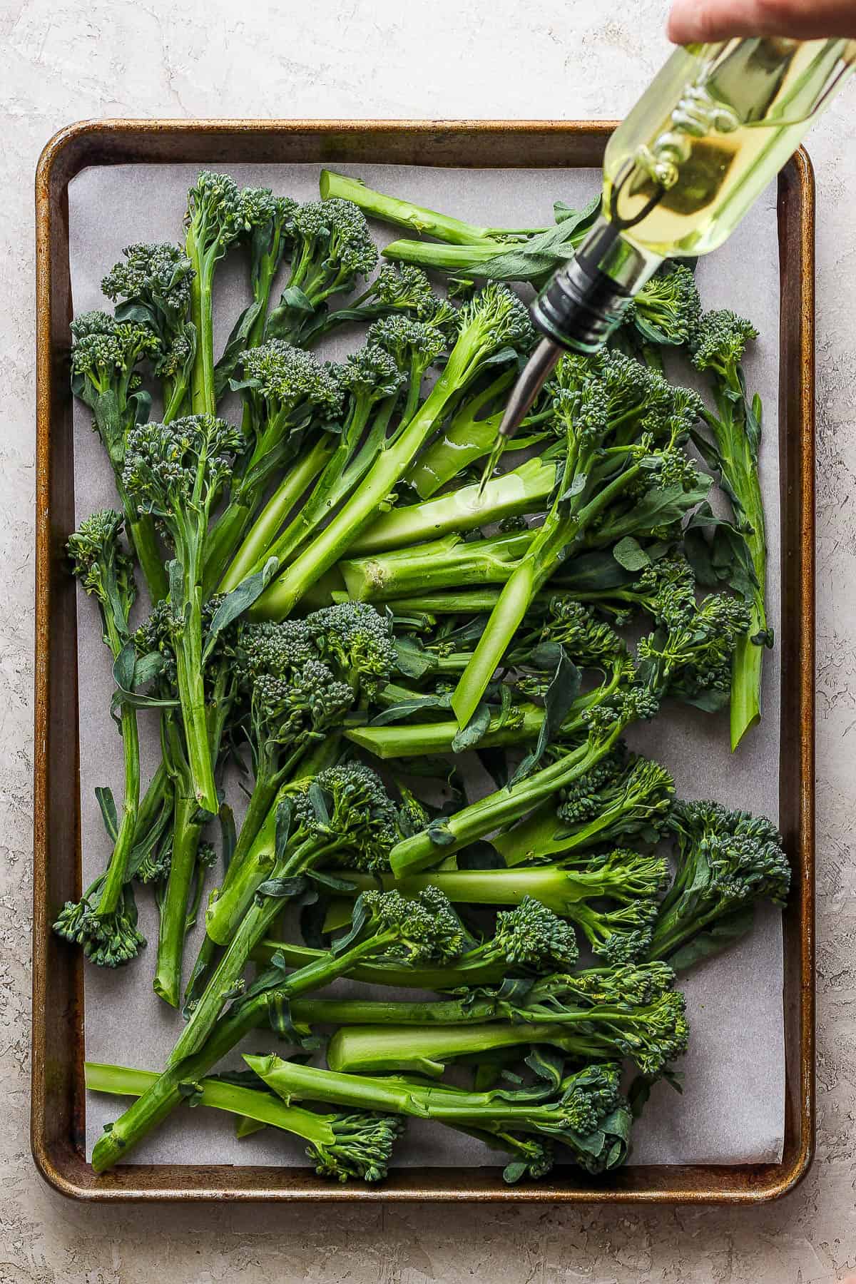 Fresh, trimmed broccolini on a parchment-lined baking sheet with olive oil being drizzled on top.