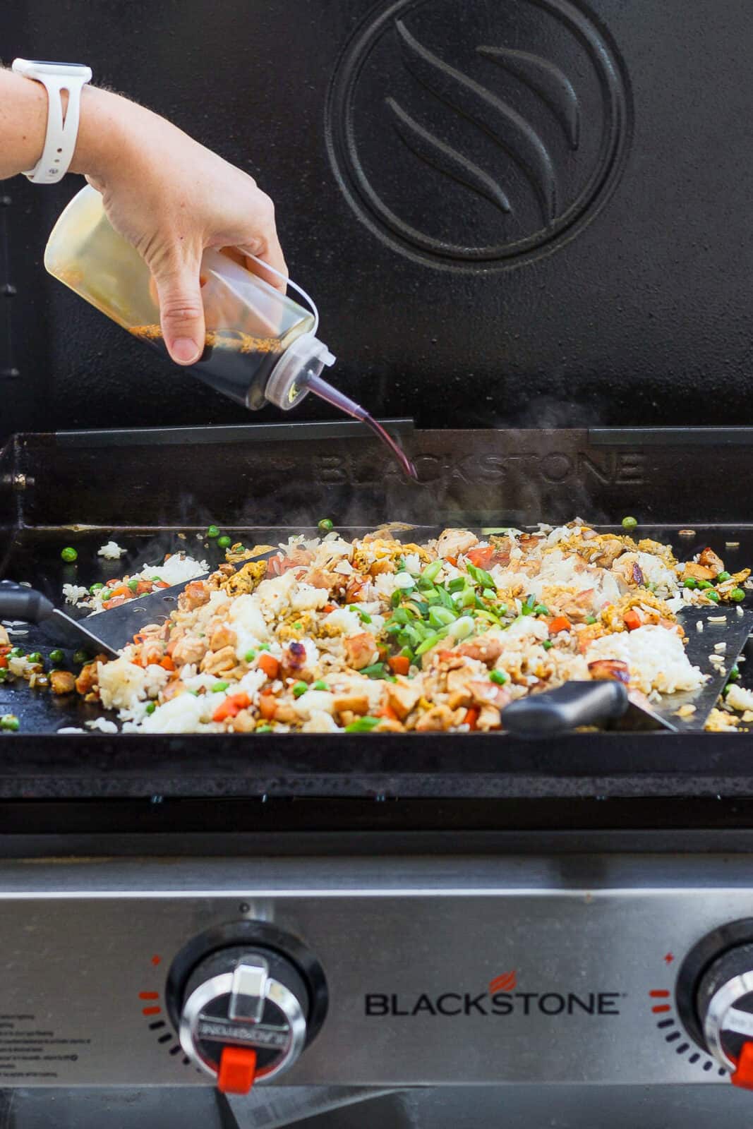 A hand pouring soy sauce out of a squirt bottle in to fried rice on the Blackstone griddle.