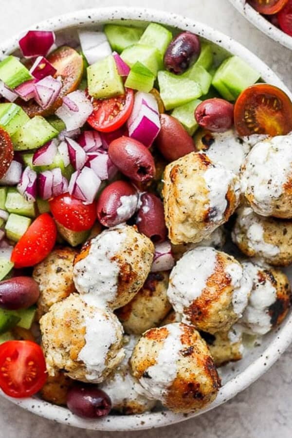 A bowl of greek chicken meatballs with tzatziki sauce and a greek cucumber salad.